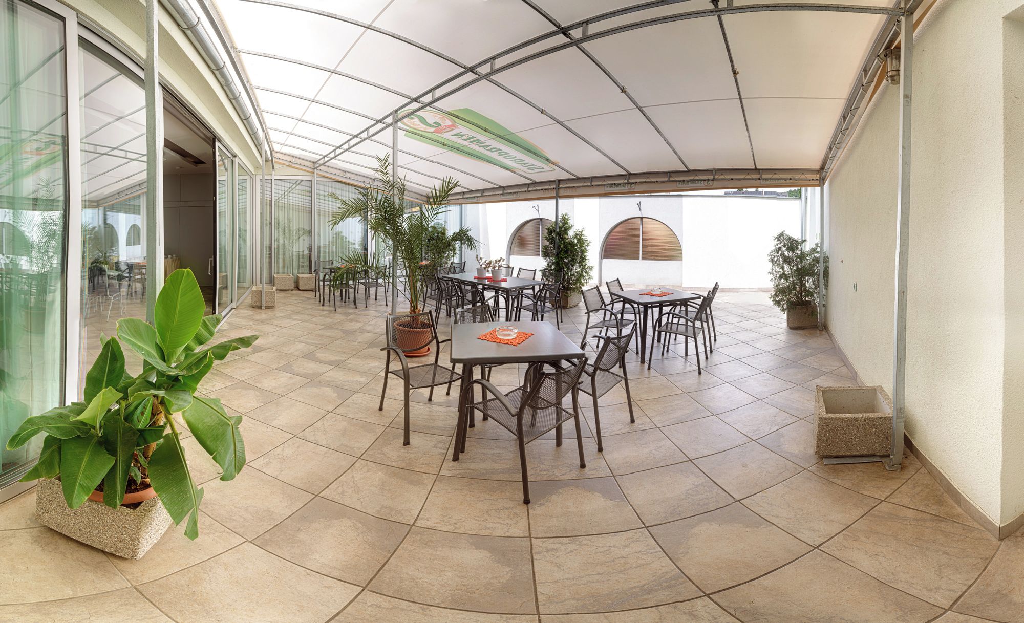 Une terrasse extérieure couverte, dotée de plusieurs tables et chaises, entourée de plantes en pot et d’un mur de verre.