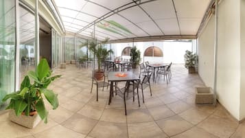 Une terrasse extérieure couverte, dotée de plusieurs tables et chaises, entourée de plantes en pot et d’un mur de verre.