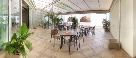 A covered outdoor patio with multiple tables and chairs, surrounded by potted plants and a glass wall.