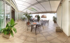 Un patio al aire libre techado con mesas y sillas, rodeado de plantas en macetas y un muro de cristal.