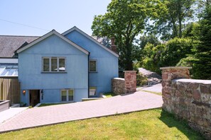 A two-story residential building with a sloped roof, a front entrance, and a paved driveway leading to a stone wall. - Cawsand Home With Garden (Saltash)