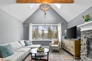 A modern living room with a stone fireplace, a sofa, a television, and a large window with a view of trees. - Charming 2BR Kisilano Duplex | Near Jericho Beach (Vancouver)