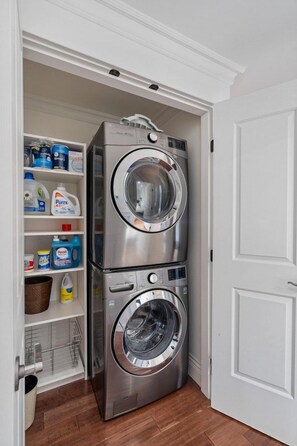 A compact laundry room with a stacked washer and dryer, shelves with laundry detergent, and a door. - Upscale West Vancouver Estate with Movie Room (West Vancouver)