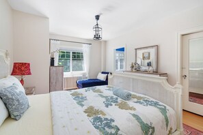 Ein Schlafzimmer mit einem großen Bett, einem Fenster mit Vorhängen, einem Heizkörper, einem blauen Stuhl und einem Spiegel an der Wand.