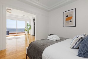 A hotel room with a bed, a view of the sea through large windows, and a framed abstract artwork on the wall. - Waterfront Home on Marine Dr | Steps to the Beach (White Rock)
