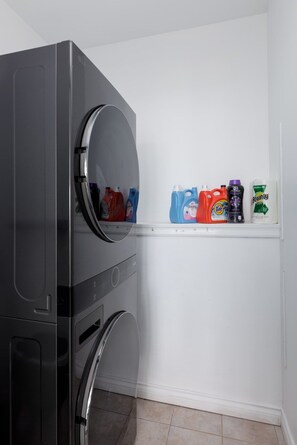A laundry area with a washing machine and a shelf with various cleaning products. - Waterfront Home on Marine Dr | Steps to the Beach (White Rock)