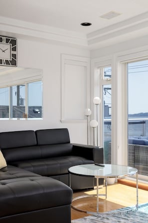 A modern living room with a black leather sofa, a glass coffee table, and a view of the water through large windows. - Waterfront Home on Marine Dr | Steps to the Beach (White Rock)
