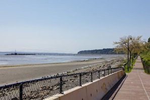 A coastal walkway with a fence, a pier in the distance, and a clear sky. - Waterfront Home on Marine Dr | Steps to the Beach (White Rock)