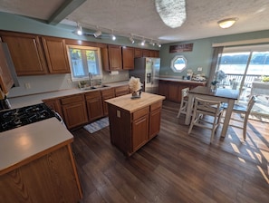 A kitchen with wooden cabinets, a central island, a stove, and a dining area with a table and chairs. - Dogs Stay Free! Fenced Lakefront House with Deck (Delton)