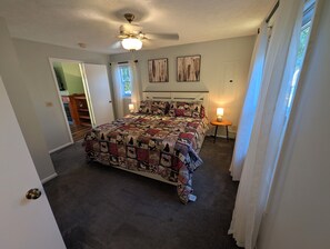 A bedroom with a bed, a ceiling fan, a nightstand with a lamp, and a window with curtains. - Dogs Stay Free! Fenced Lakefront House with Deck (Delton)