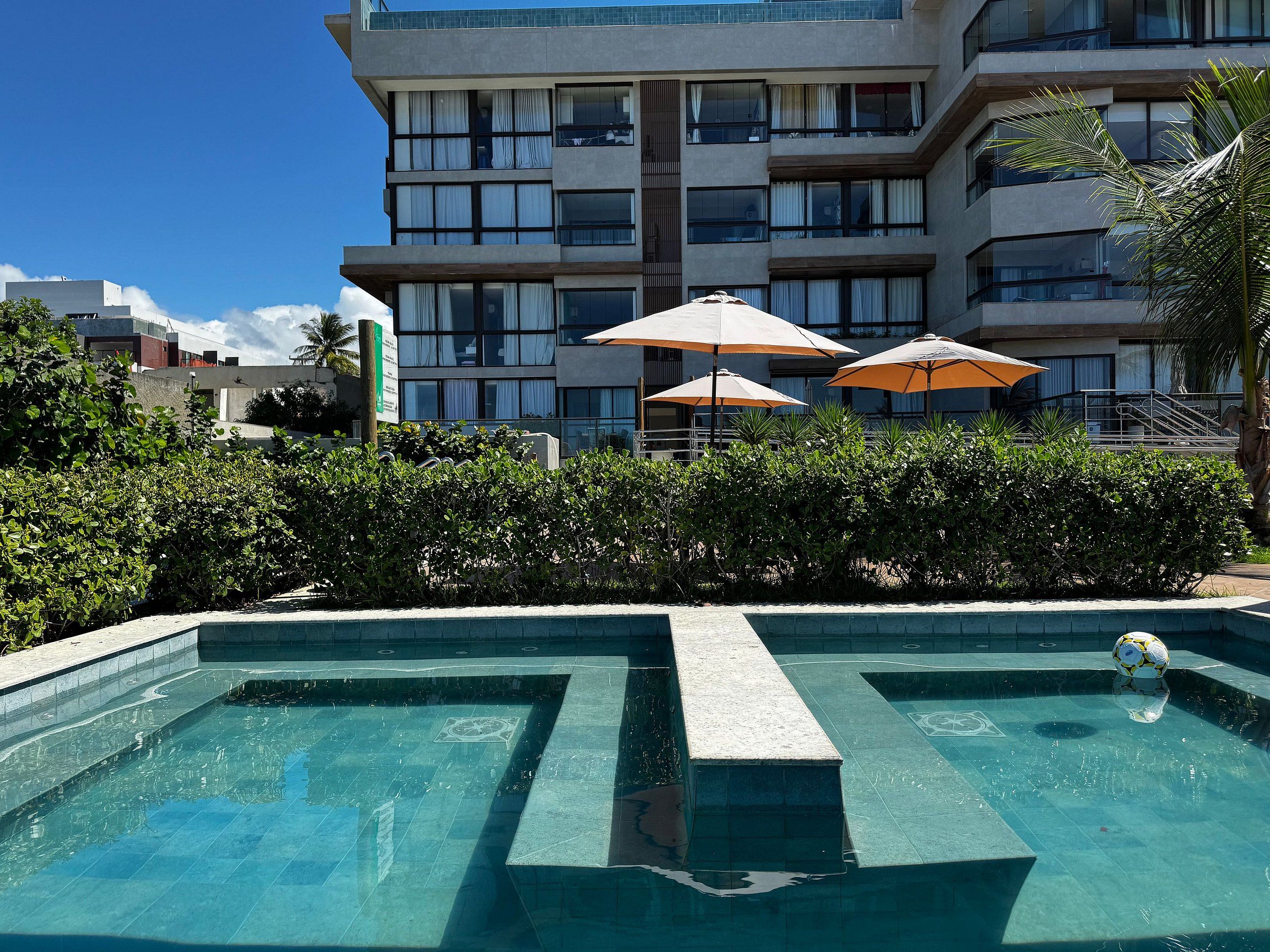 A swimming pool with a geometric design, surrounded by a hedge and umbrellas, with a multi-story building in the background.