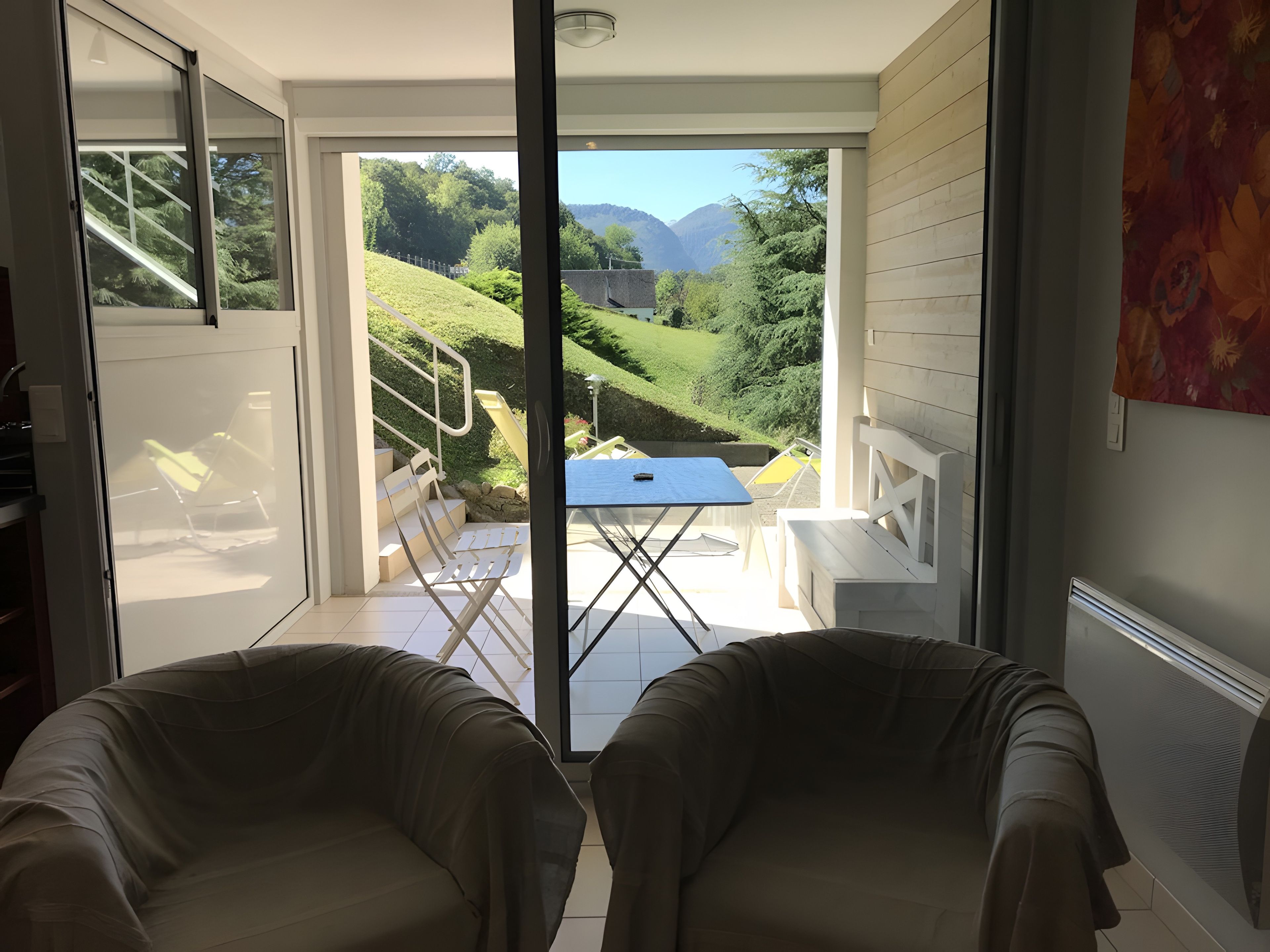 Una habitación con puerta de vidrio corredera que da a un espacio exterior con mesa y sillas, una escalera y vistas a un paisaje verde.