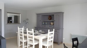 Un comedor con una mesa y sillas blancas, un armario gris con estantes y un espejo en la pared.