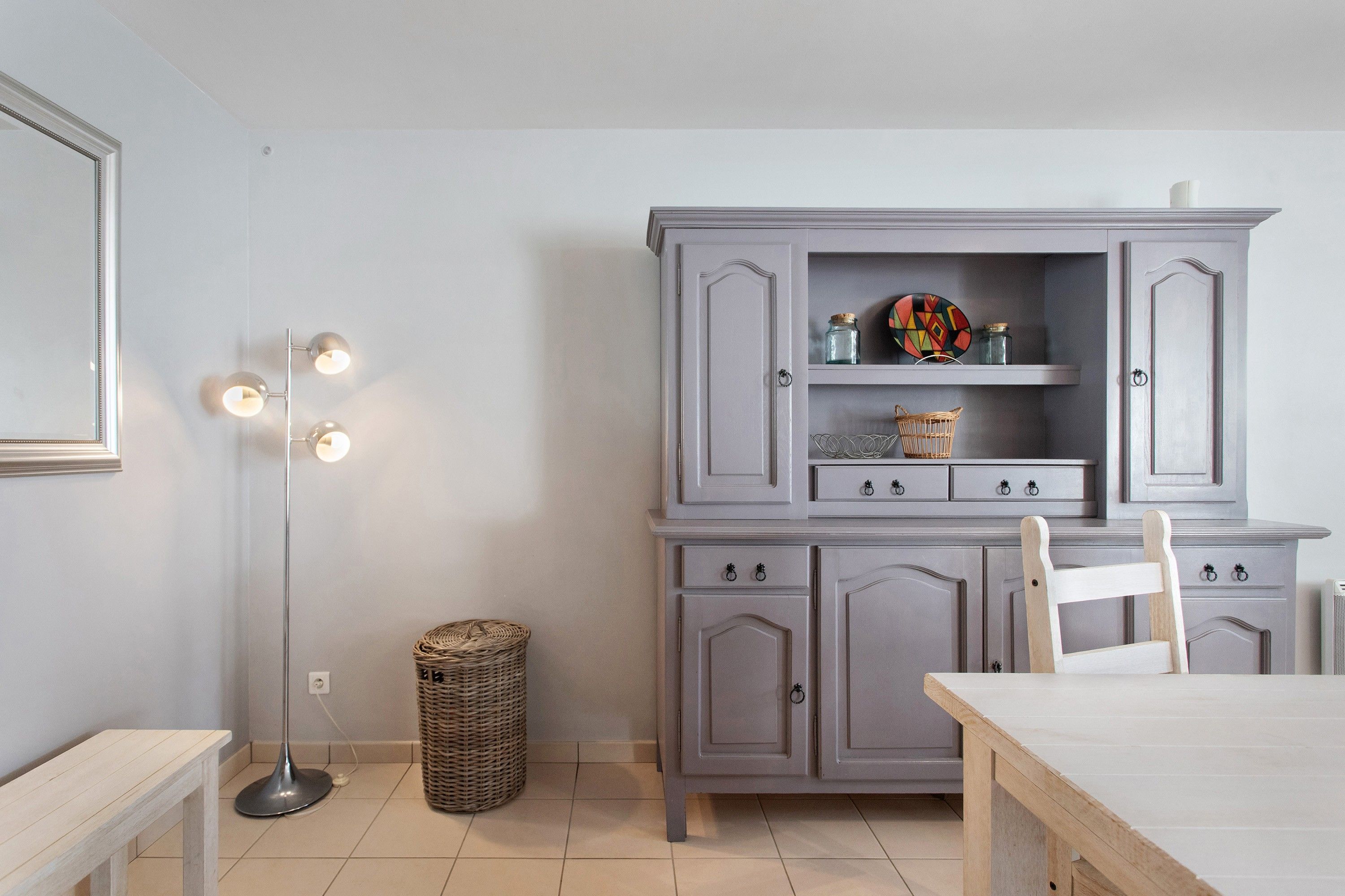 Une pièce avec un meuble gris, un panier en osier, une lampe sur pied et une table en bois.