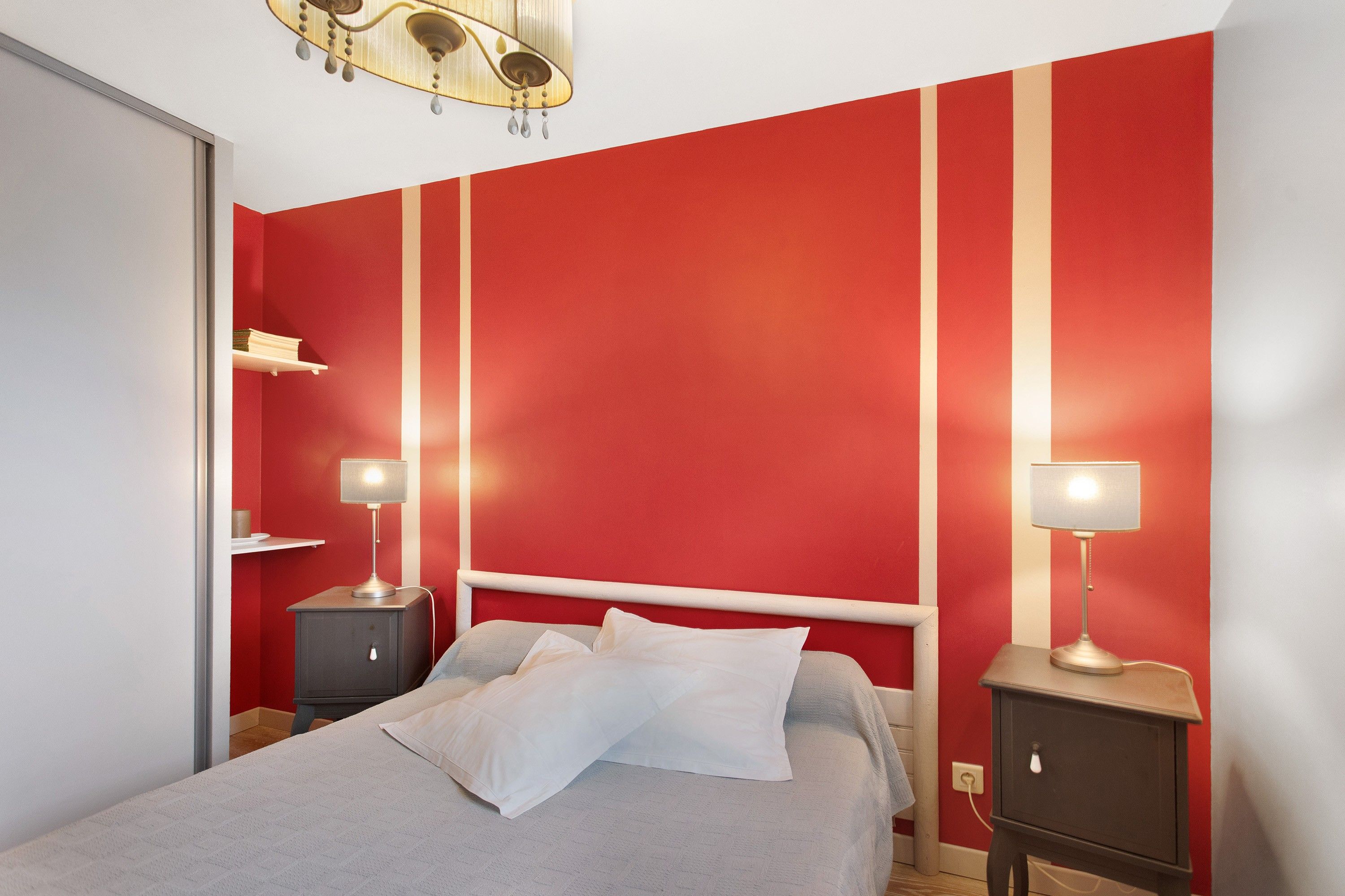 A bedroom with a bed, two bedside tables with lamps, a wardrobe, and a red striped wall.