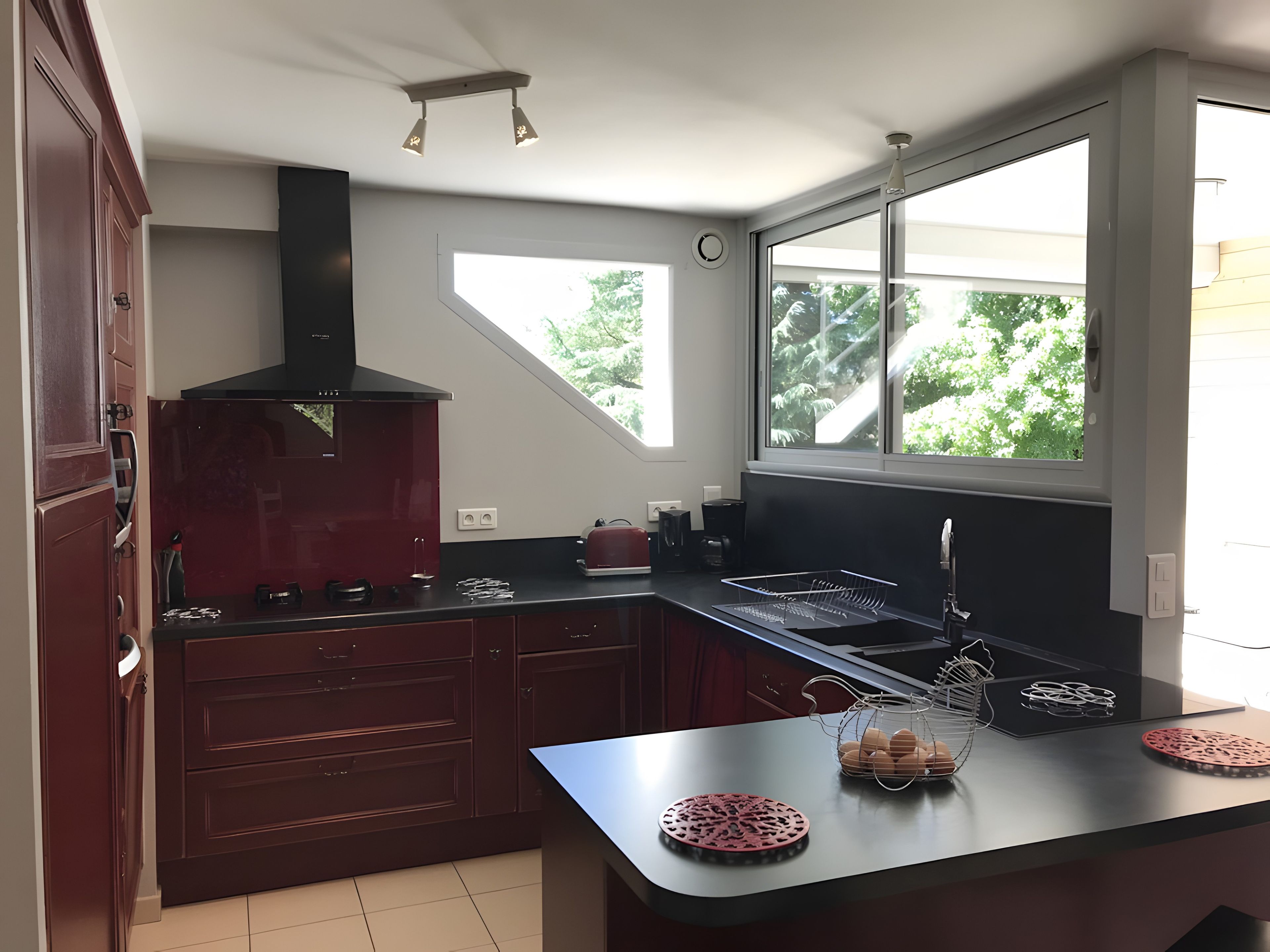 Een moderne keuken met donkere kastjes, een zwarte afzuigkap, een raam met uitzicht op groen, en een aanrecht met een koffiezetapparaat en een mand.