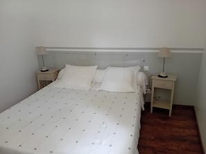Un lit simple avec une couette et des oreillers blancs, deux tables de chevet avec des lampes et un plancher en bois.