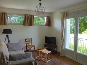 Un salon comprenant un canapé, un fauteuil, une petite table, un téléviseur et une lampe. Il y a des fenêtres avec des rideaux, et de l’extérieur, on peut voir de la verdure.