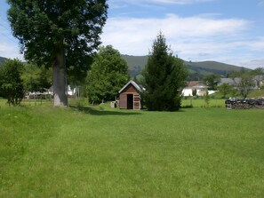 A small wooden cabin in a grassy field with trees and houses in the background.