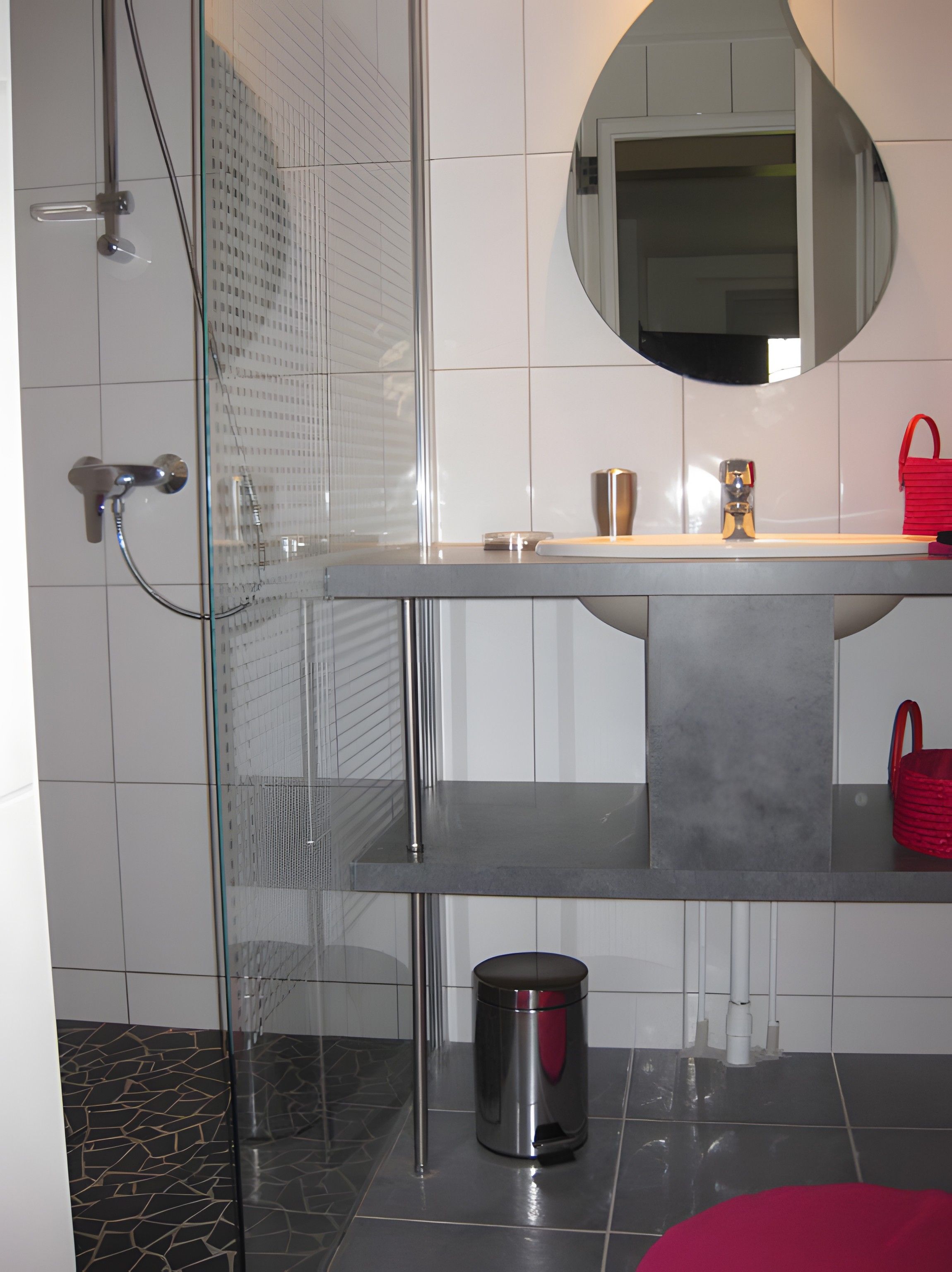 Une salle de bain moderne avec une douche à parois de verre, un miroir rond, un lavabo en acier inoxydable et un sac à main rouge posé sur le comptoir.