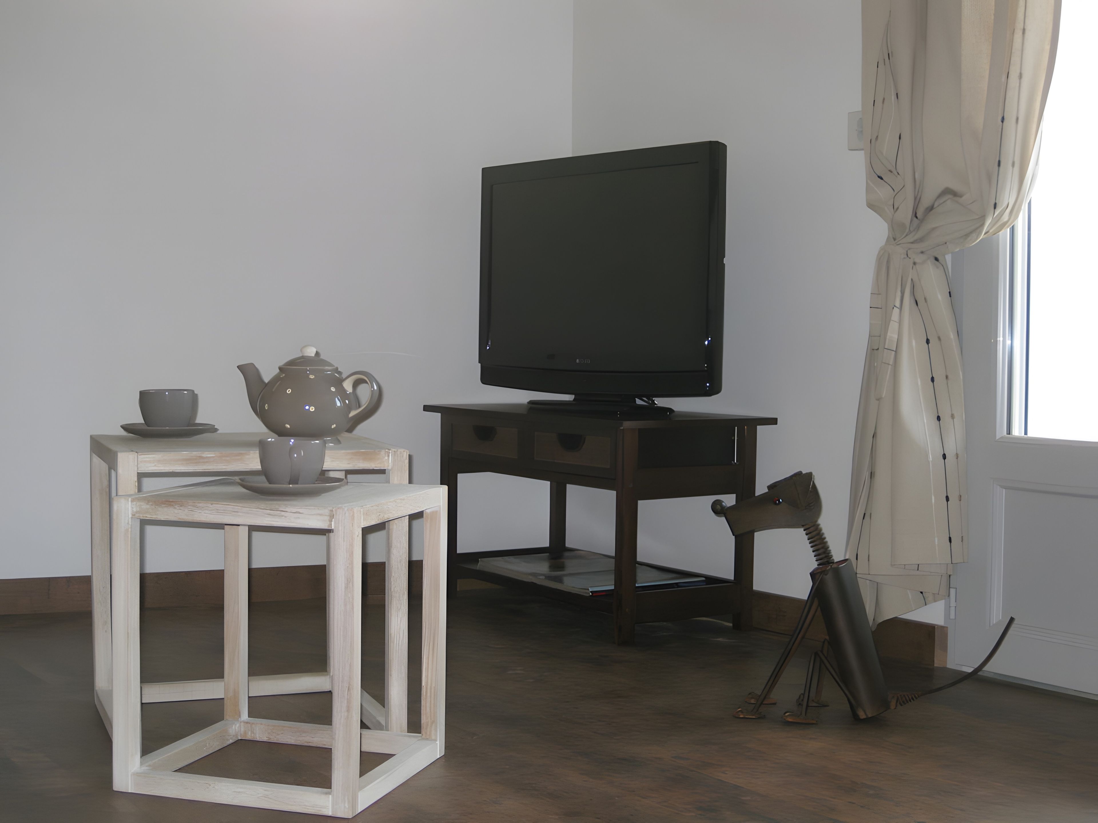 Une pièce avec un téléviseur posé sur un meuble en bois, une petite table avec une théière et une tasse, et une fenêtre avec des rideaux.