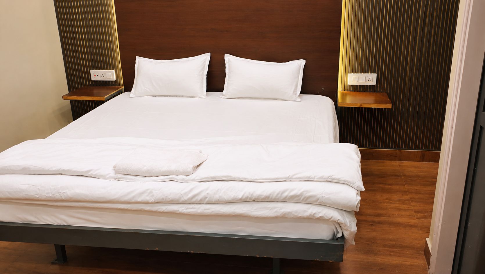 Cama bem arrumada com lençóis brancos, cabeceira de madeira e duas mesas de cabeceira com abajures.