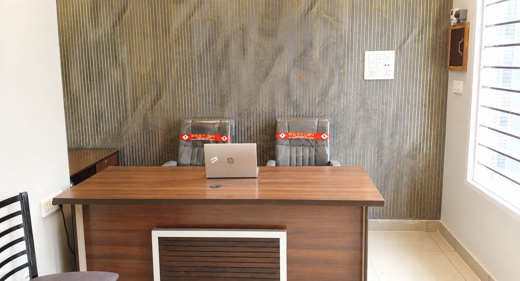A conference room with a wooden table, two chairs, a laptop, and a patterned wall.