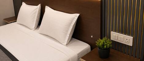 A neatly made bed with white bedding and pillows, a bedside table with a potted plant, and a wall-mounted electrical outlet.
