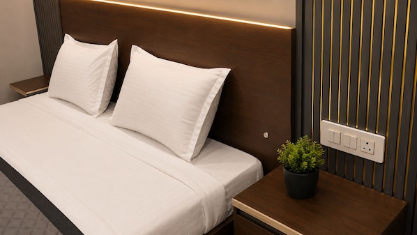 A neatly made bed with white bedding and pillows, a bedside table with a potted plant, and a wall-mounted electrical outlet.