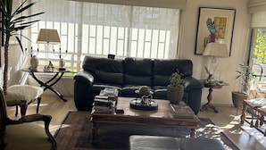 Un salon avec un canapé en cuir noir, une table basse en bois, un tapis à motifs et une grande fenêtre avec des stores.