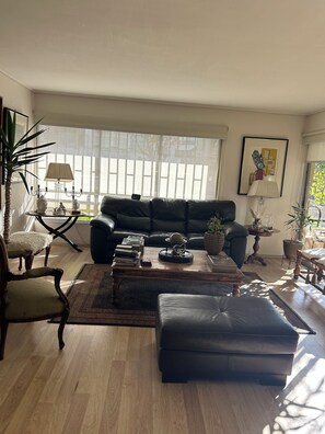 Een woonkamer met een zwarte leren bank, een houten salontafel, een tapijt met dessin en een groot raam met zonwering.