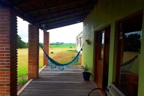 A wooden porch with a hammock, a potted plant, and a view of a green field.