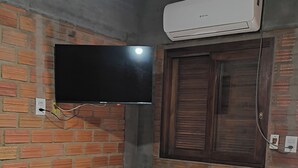 A room with a brick wall, a flat-screen TV mounted above a wooden table and chair, an air conditioning unit, and a bed with patterned bedding.