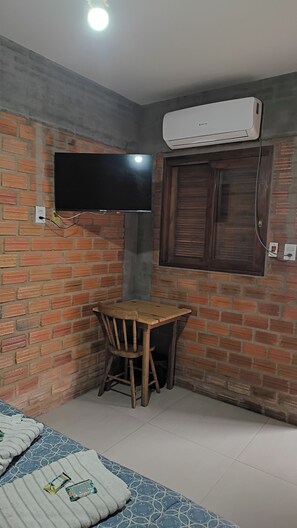 A room with a brick wall, a flat-screen TV mounted above a wooden table and chair, an air conditioning unit, and a bed with patterned bedding.
