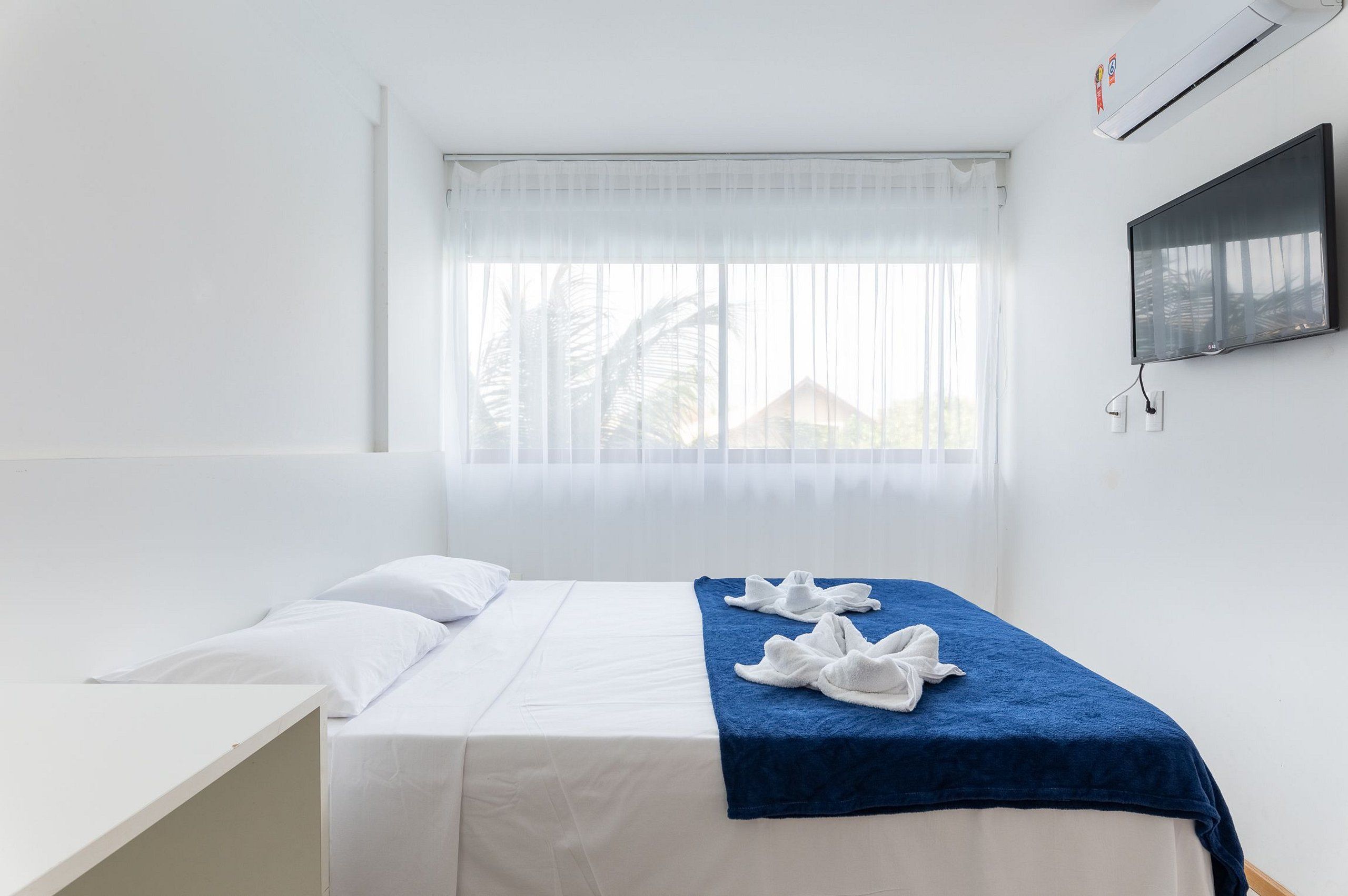 A single bed with white bedding and a blue blanket, a white bedside table, a mounted TV, and a window with sheer curtains.