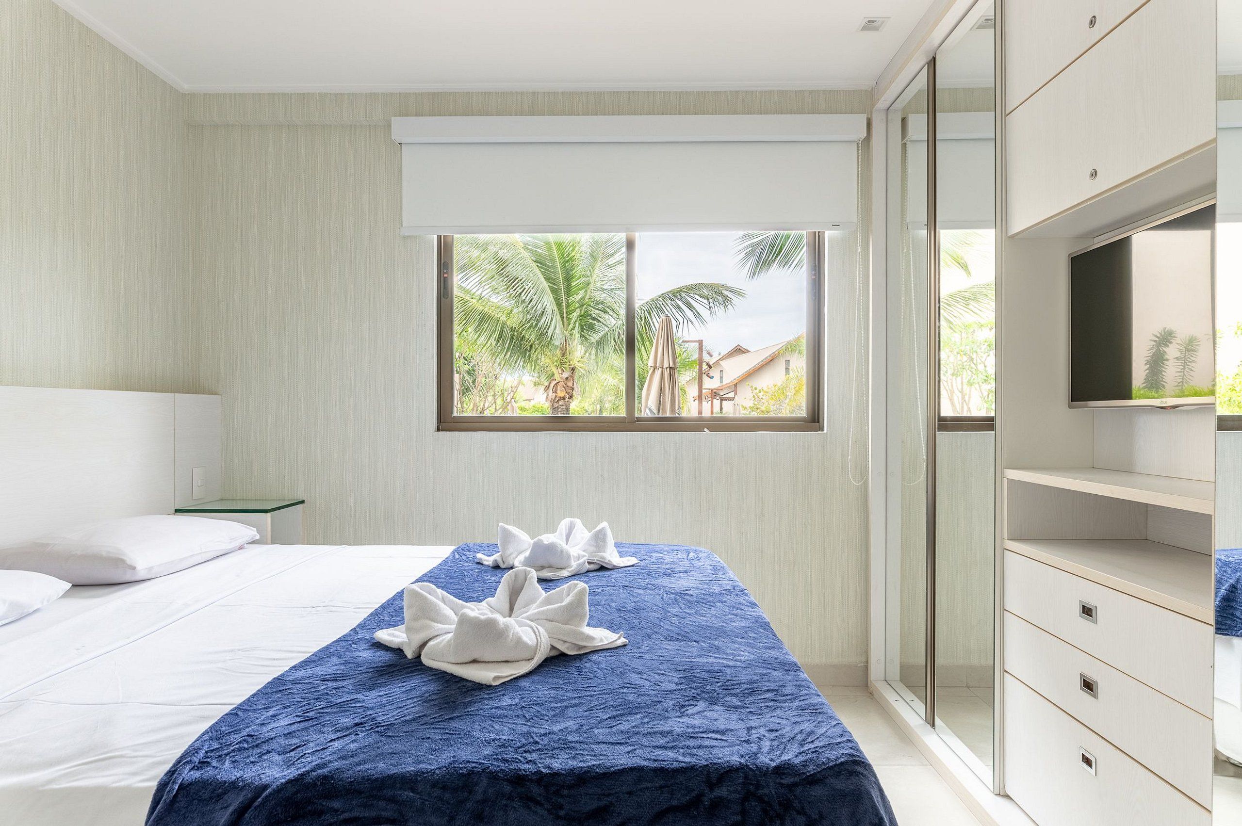 A hotel room with a bed, a window with a view of palm trees, and a wardrobe.