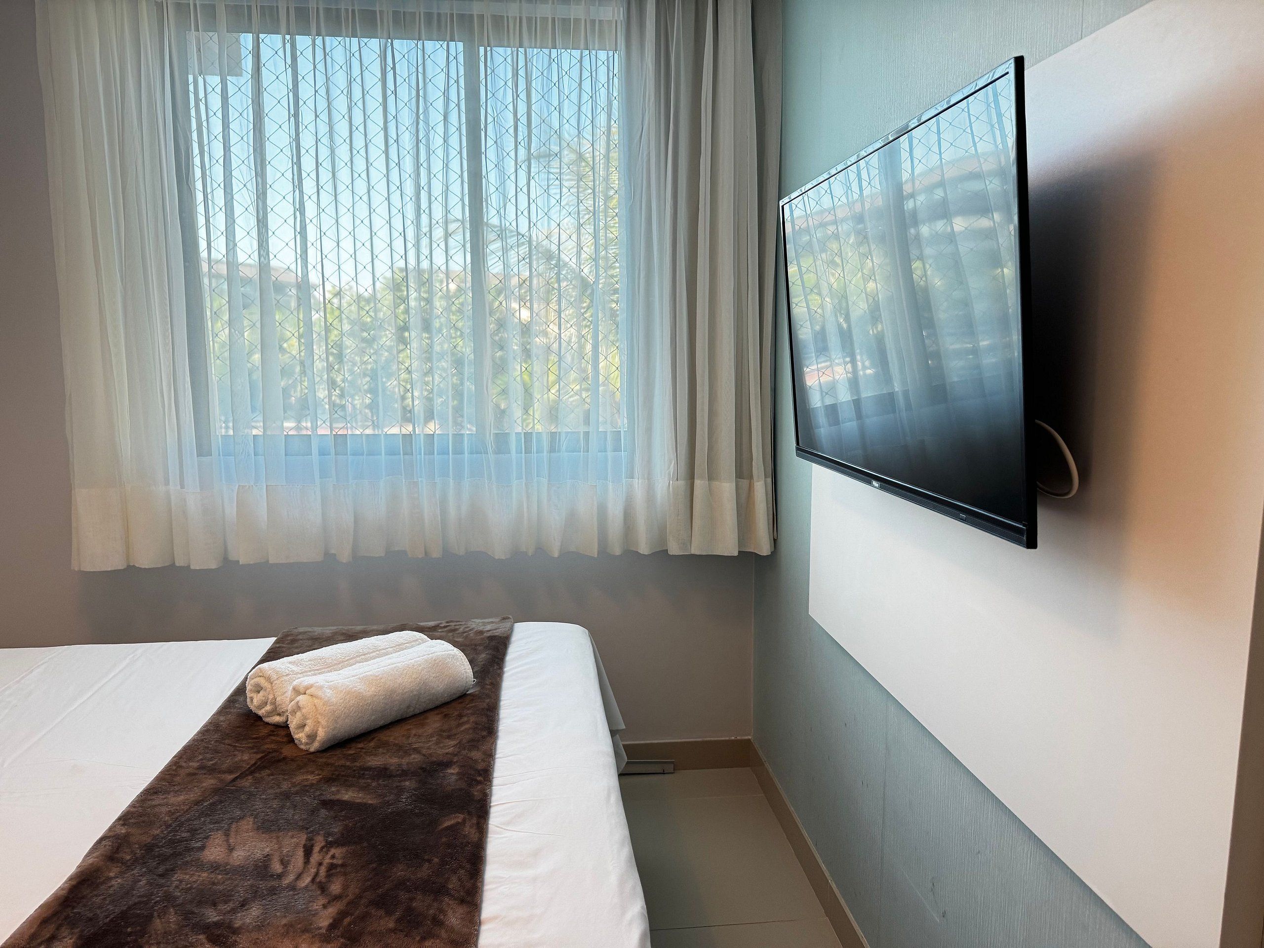 Una habitación de hotel con cama, televisor montado en la pared y ventana con cortinas traslúcidas.
