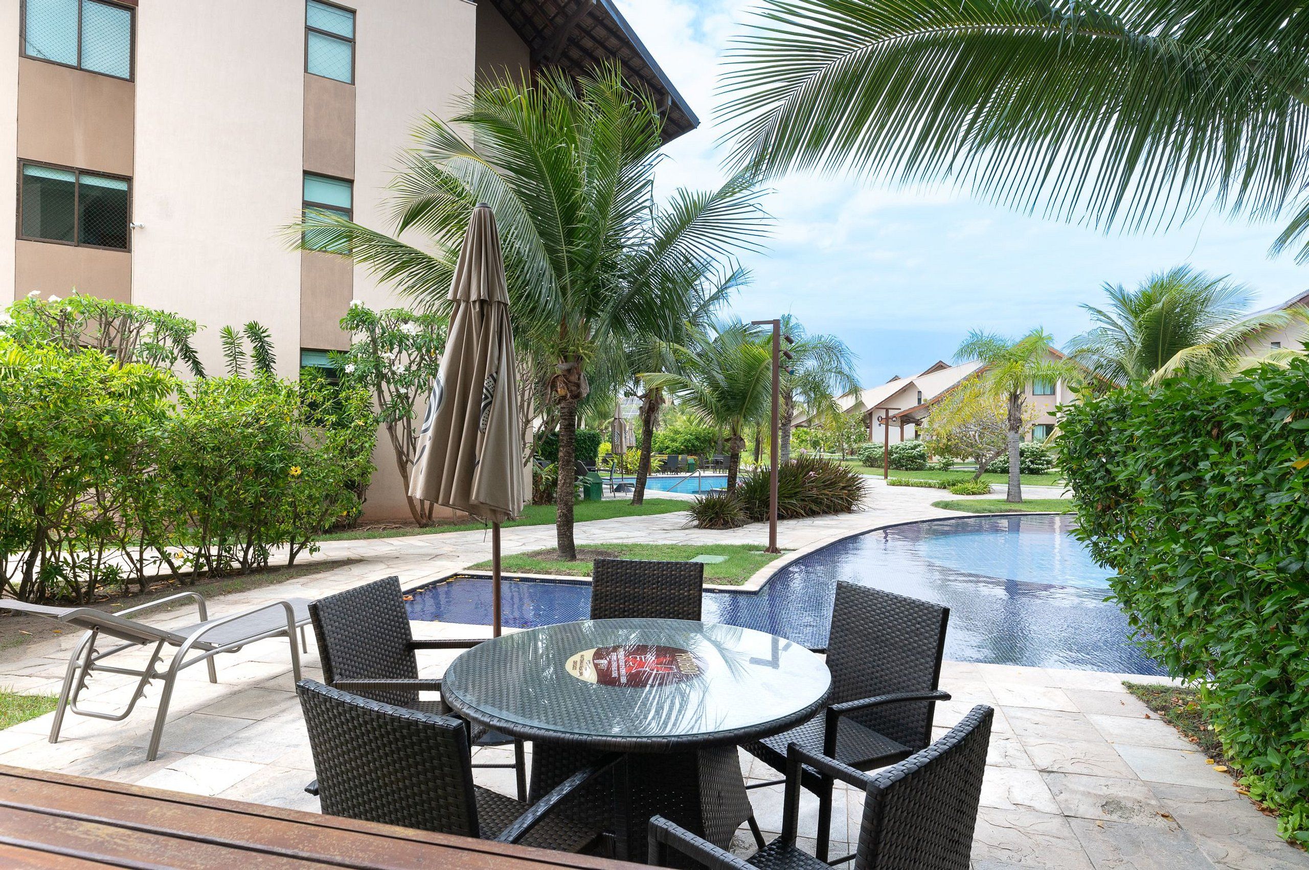 A poolside area with a round table and chairs, surrounded by palm trees and a swimming pool.