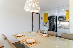 Ein Essbereich mit einem Glastisch, gedeckt für vier Personen, eine moderne Küche mit Kühlschrank und Mikrowelle sowie eine Pendelleuchte über dem Tisch.