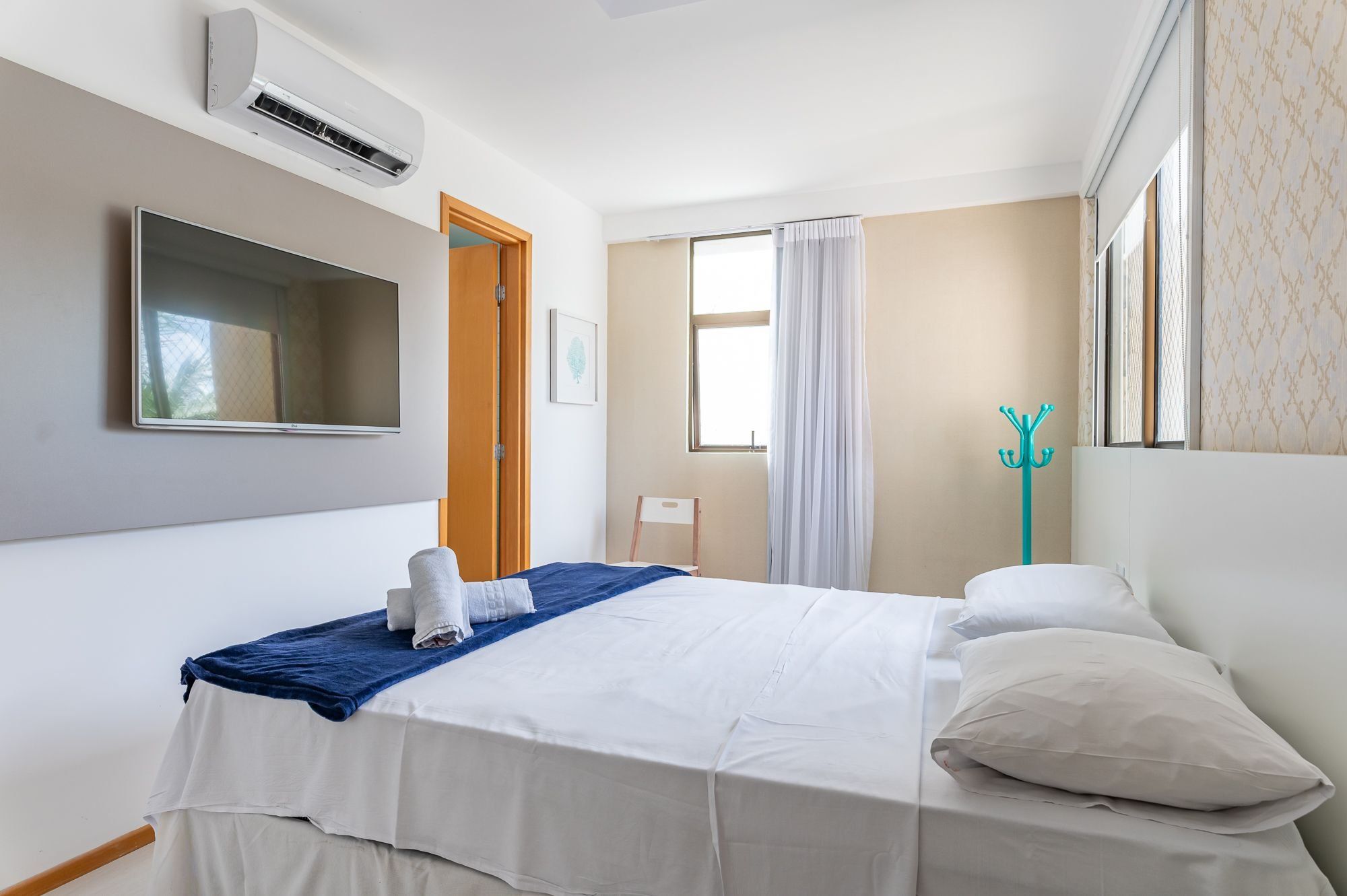 A hotel room with a bed, a TV, an air conditioning unit, a window with curtains, and a coat rack.