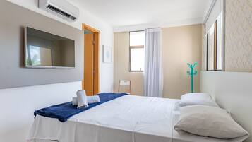 A hotel room with a bed, a TV, an air conditioning unit, a window with curtains, and a coat rack.