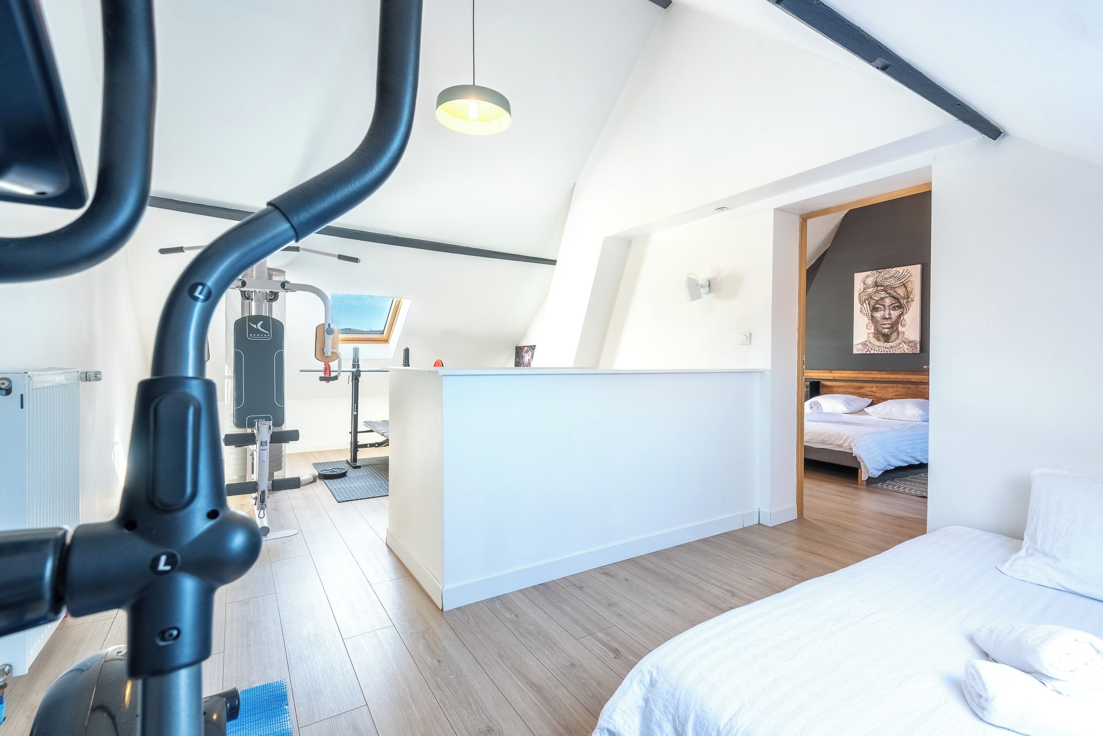 Un dormitorio moderno con cama, una zona de ejercicios con equipamiento y vistas al cielo a través de un tragaluz.