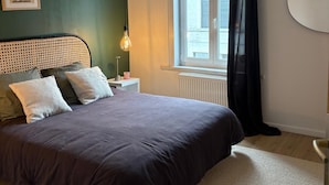 A bedroom with a bed, a nightstand, a lamp, a window with curtains, and a radiator. - Vrbo Property (Croix)