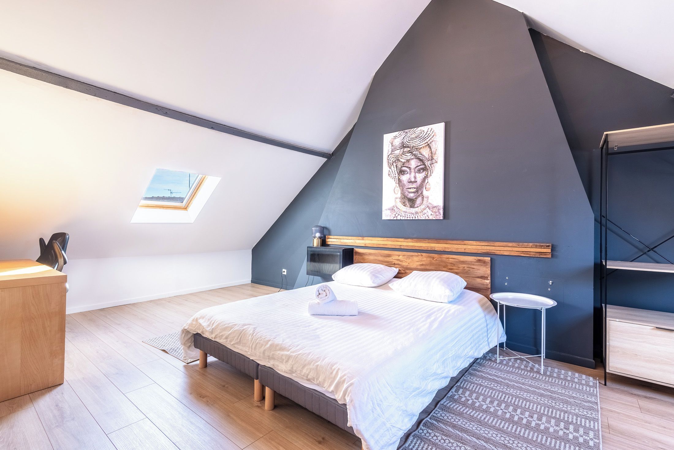 A bedroom with a sloped ceiling, a bed with white bedding, a bedside table, a small round table, and a wall-mounted shelf.