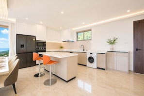 A modern kitchen with a central island, bar stools, a built-in oven, a washing machine, and a view of the outdoors.