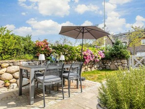 Un patio al aire libre con una mesa, sillas, un gran paraguas y una pared de piedra con plantas florecientes.
