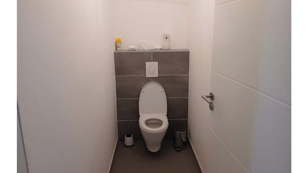 A small, narrow bathroom with a toilet, a trash can, and a toilet paper holder on the wall. - Vrbo Property (Le Boulou)