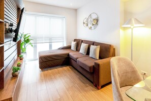 A modern living room with a brown sectional sofa, a glass coffee table, a floor lamp, and a large window providing natural light. - 90m² apartment with parking, just steps from Kennedy Park. (Miraflores)