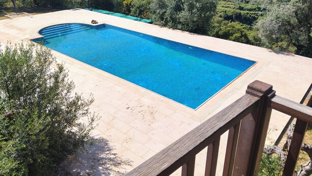 A rectangular swimming pool with a wooden deck surrounding it, set against a backdrop of a hilly landscape with trees and a distant building. - Isolated luxury villa with pool, 30 mins to sea, 2 km to village shops tavernas (messenia)