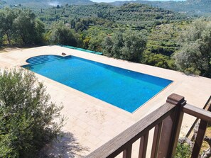 Una piscina rectangular con una terraza de madera que la rodea, con un paisaje montañoso de árboles y un edificio a la distancia como fondo.